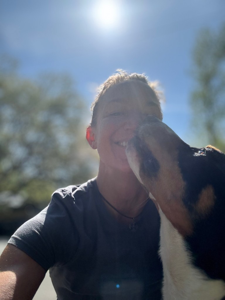 A smiling person with a dog nuzzling their face beneath a bright blue sky.