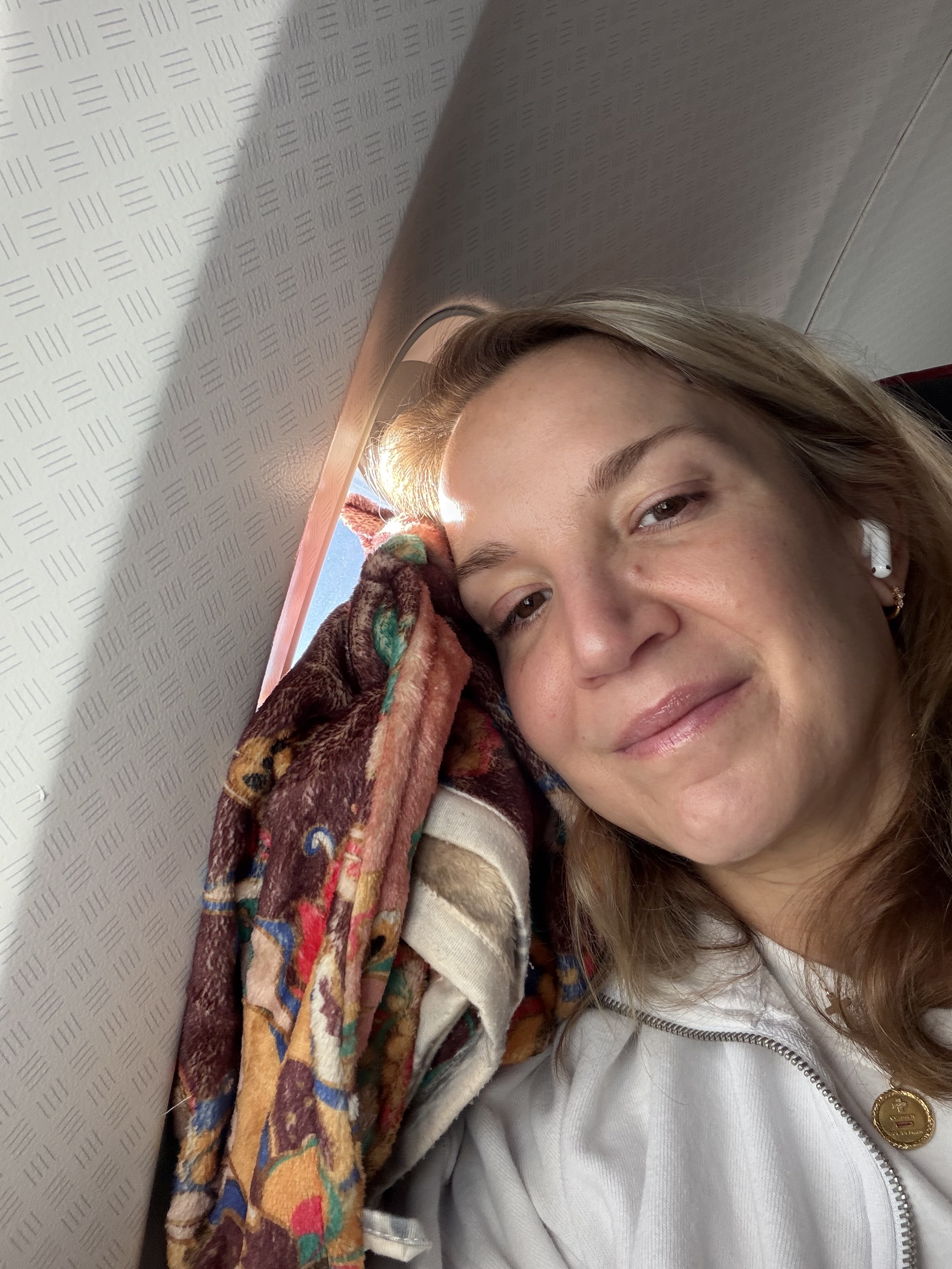 Woman wearing an earbud, resting beside an airplane window with a colorful blanket.