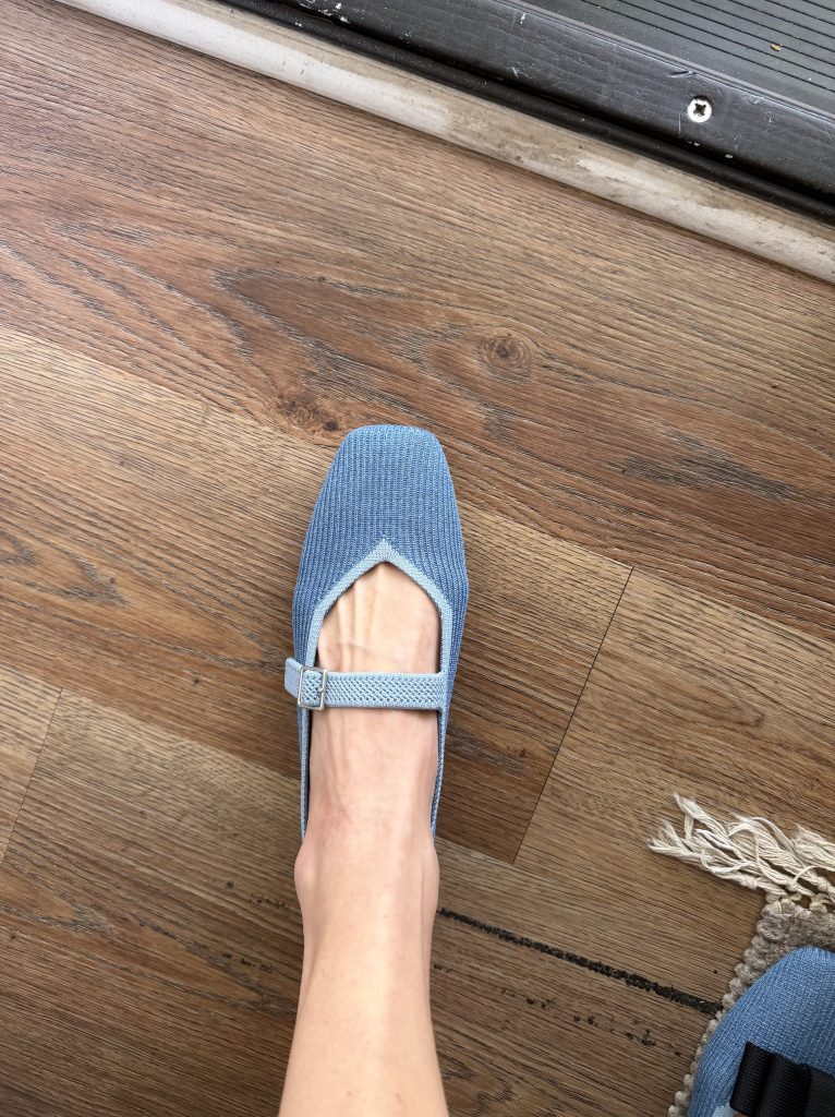 A blue knit Mary Jane shoe with a buckle strap on a wooden floor.