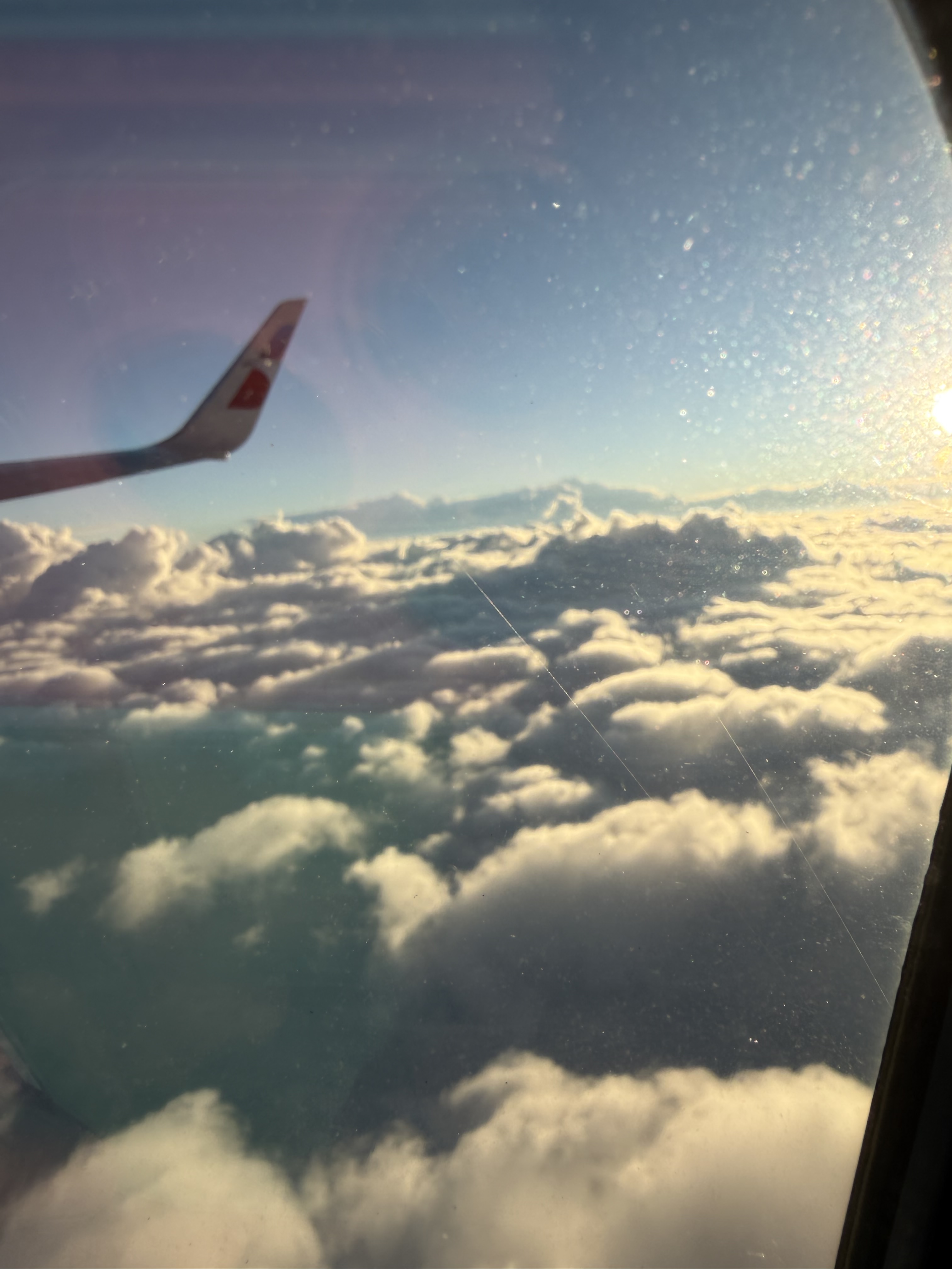 Airplane wing above sunlit clouds, seen through the window.